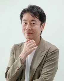 淡い背景の前で、薄茶色のジャケットを着用したミドルエイジの男性が、顔に手を添えながらカメラに向かって微笑んでいます。落ち着いた雰囲気のポートレート写真です。
