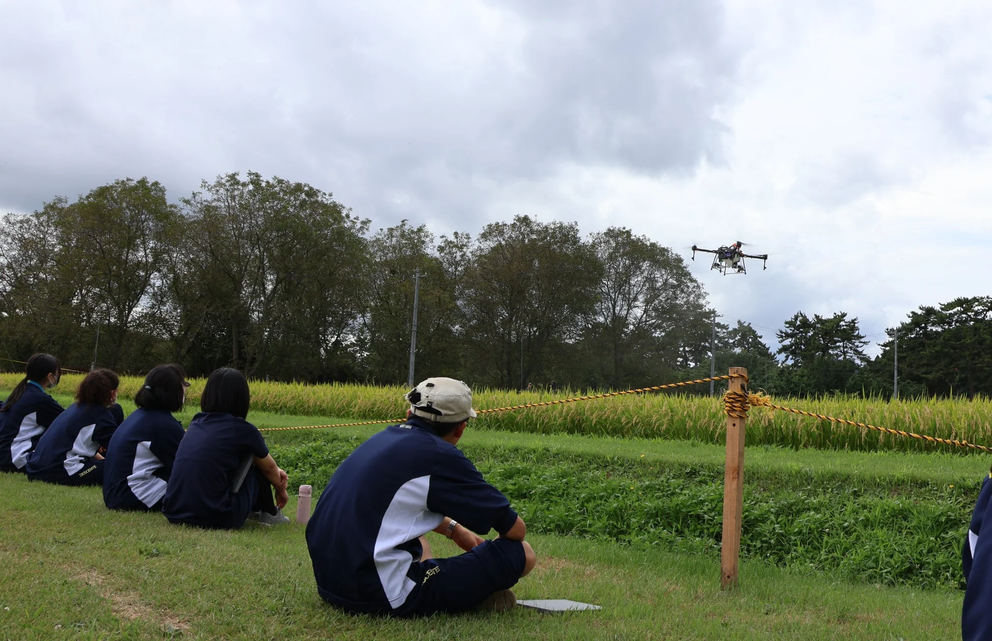 人々が芝生に座り、水田の上空を飛行するドローンを観察している様子