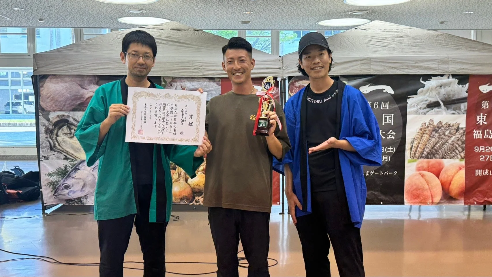 3人の男性が食品関連のイベントで表彰されている写真です。中央の男性はトロフィーを持ち、左の男性は賞状を持っています。背景には、魚介類や桃などの食品が描かれたバナーと、大会情報が掲示されています。