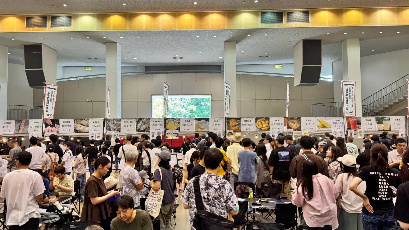 「日本の素材甲子園」というイベントで、大勢の人が賑わう屋内会場の様子。能登牛、淡路玉ねぎ、紀州うめどり、ホタテ、スイカのスムージーなど、全国各地の特産品やグルメを販売する多数の屋台が並び、来場者が食事や買い物を楽しんでいる。