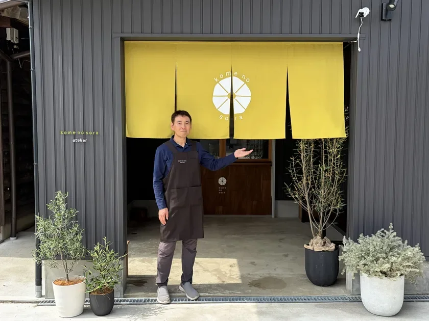 「kome no sora atelier」のモダンな店舗入口で、エプロン姿の男性が誇らしげに立っています