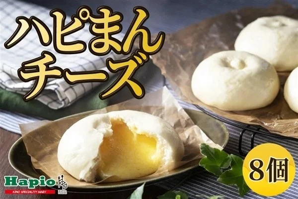 ハピオの「ハピまんチーズ」を写した画像で、白い蒸しまんじゅうが複数個並べられています。手前の1つは割れており、中からとろりとしたチーズフィリングが見えています。全体的に温かみのある雰囲気で、8個入りであることが示されています。