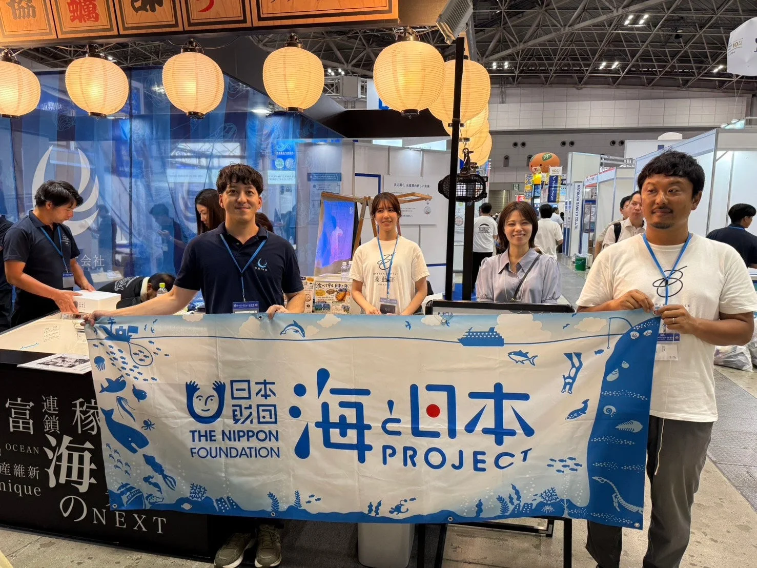 イベント会場のブースで、数人のスタッフが「日本財団 海と日本 PROJECT」と書かれたバナーを掲げています。ブースには提灯が飾られ、来場者に向けて海洋に関する取り組みを紹介している様子が伺えます。