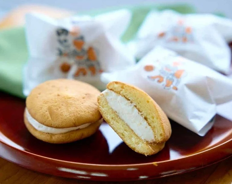 赤い皿の上に置かれた、クリームが挟まれた和菓子が写っています。一つは半分に切られており、白いクリームの層が見えています。背景には個別に包装された同じようなお菓子がいくつか並んでいます。