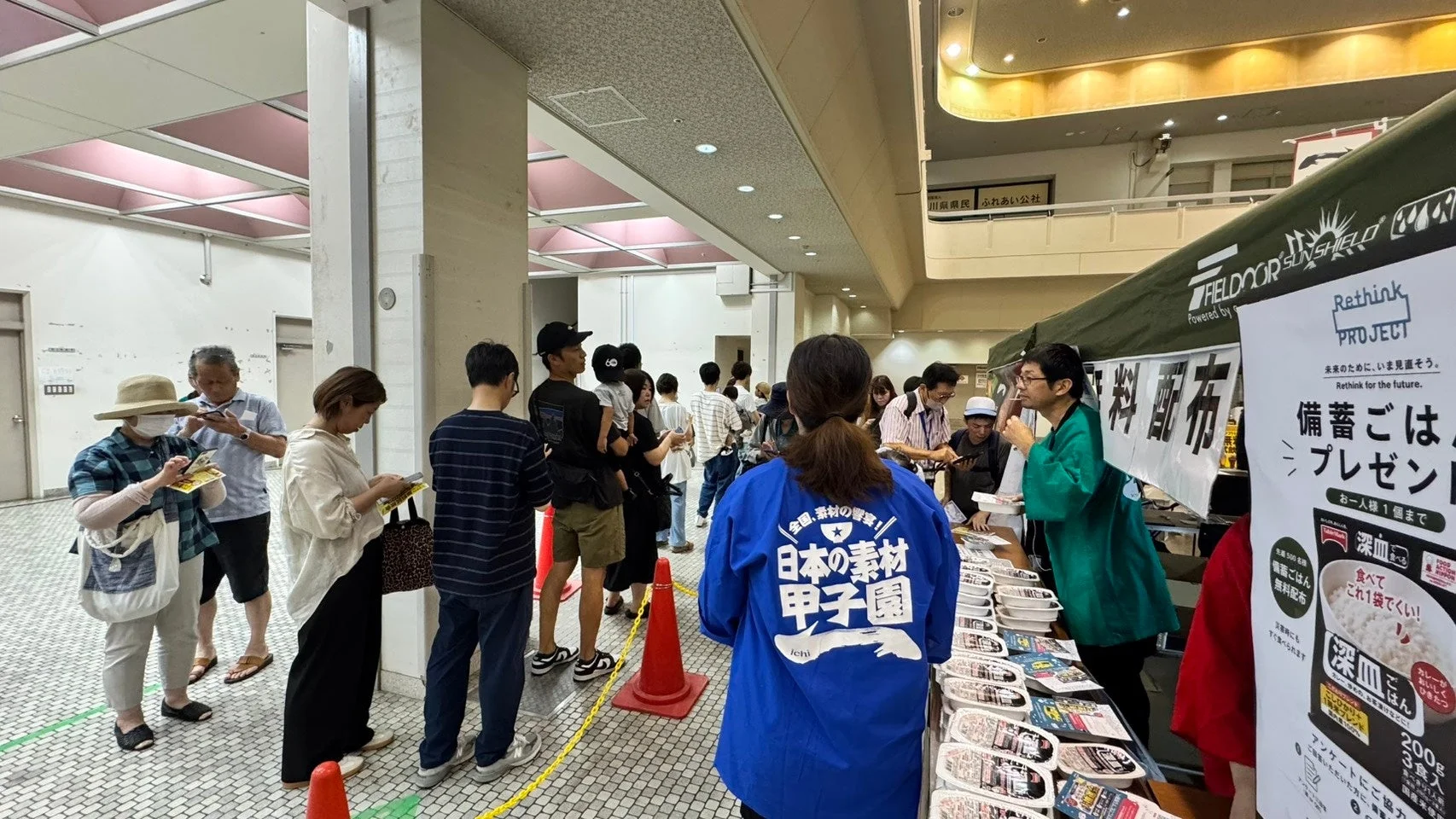 屋内で開催されているイベントで、多くの人々が「Rethink PROJECT」による備蓄ごはんの無料配布に並んでいます。「日本の素材 甲子園」と書かれた青いジャケットを着たスタッフが対応しており、防災意識を高めるための活動のようです。