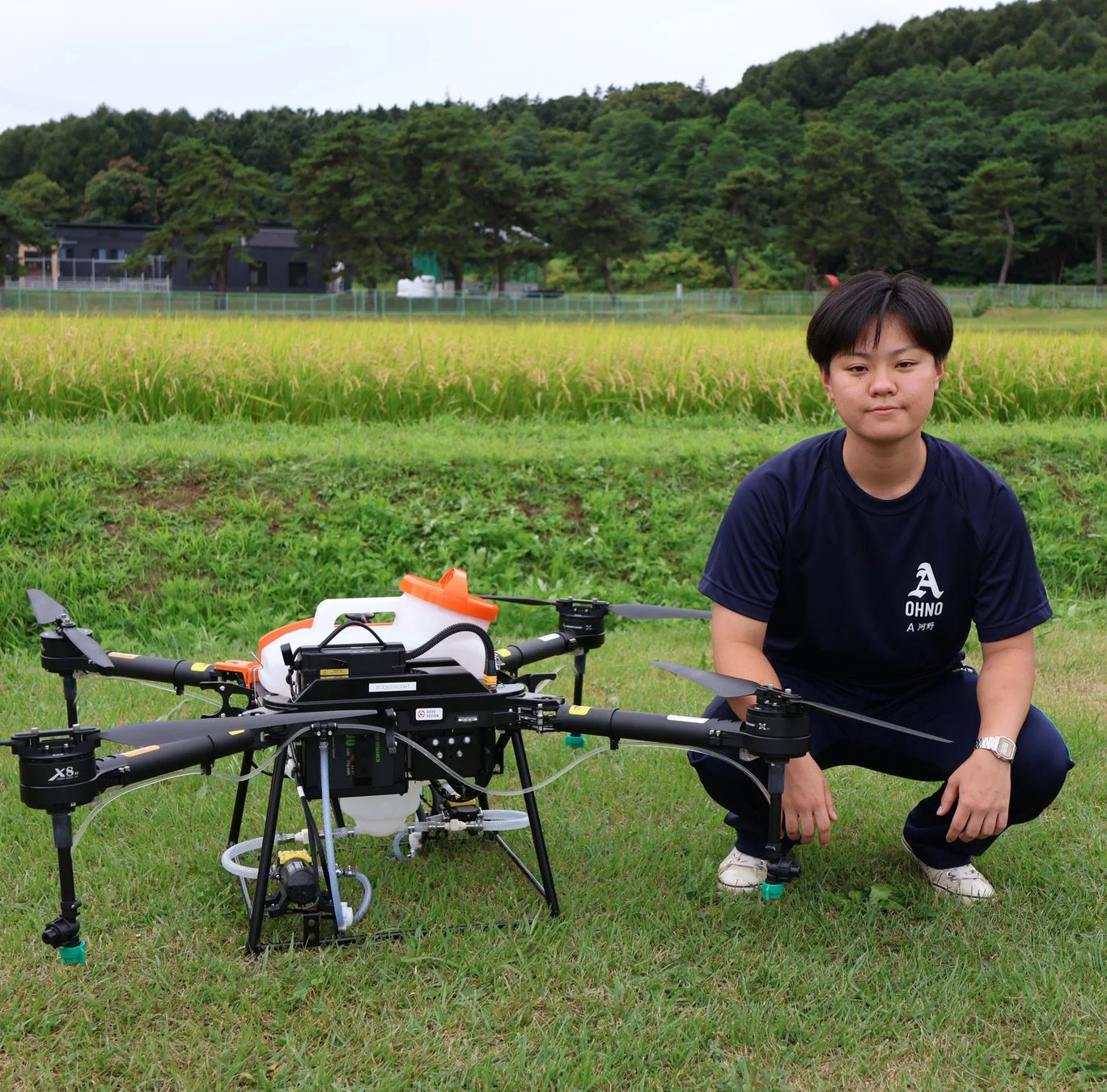 水田を背景に、農業用ドローンの横でしゃがむ人物