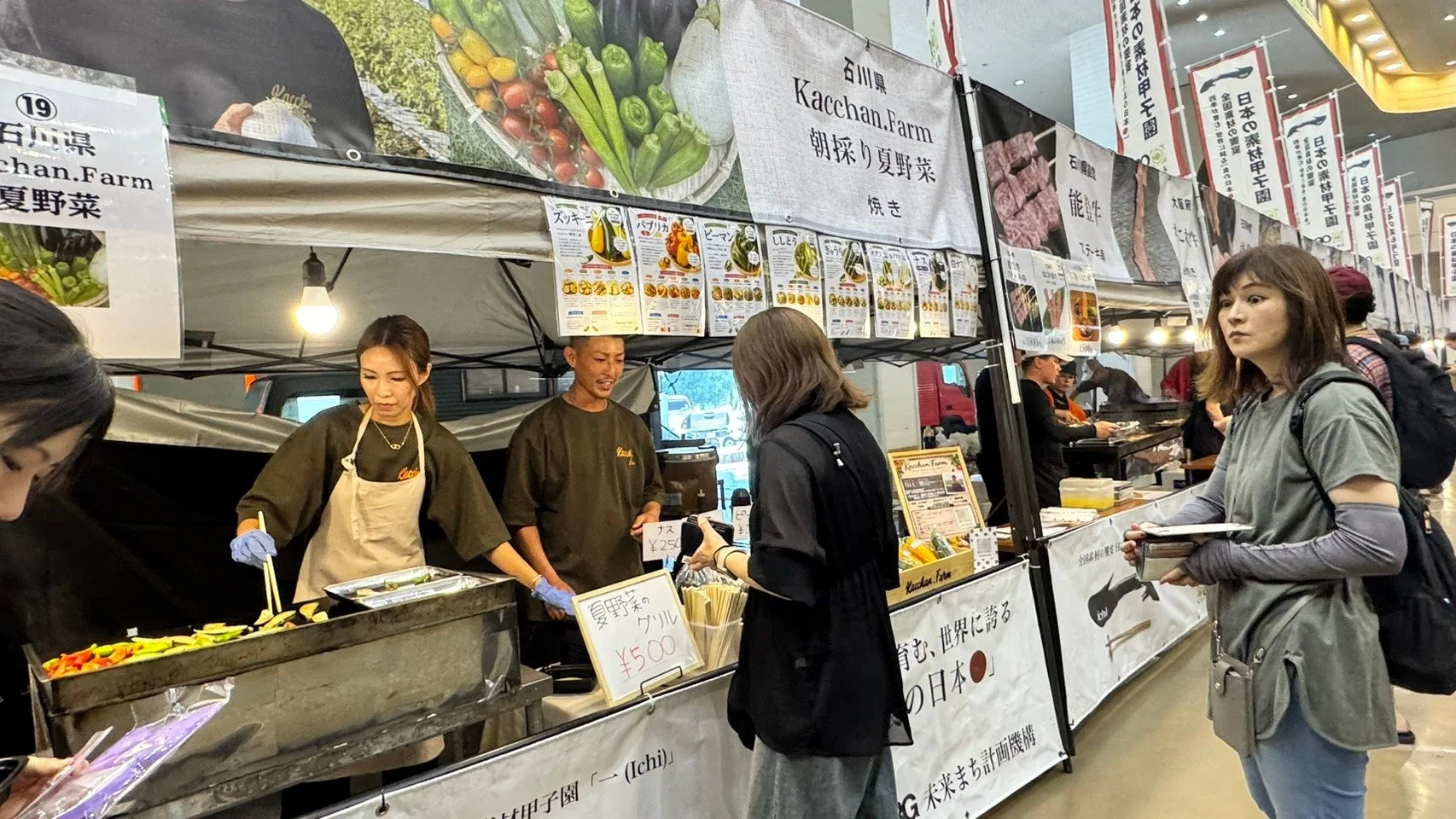 屋内のイベント会場で、石川県の「Kacchan.Farm」が朝採り夏野菜のグリルを販売している様子です。女性スタッフが調理し、男性スタッフが接客しています。多くの来場者がいて、他の屋台のぼりも見えます。