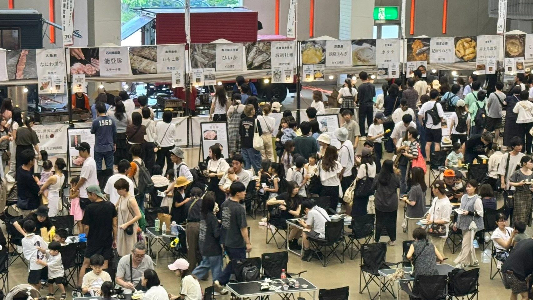 多くの人々で賑わう屋内イベントの様子です。様々な地域の特産品を販売するフードブースが並び、多くの来場者が食事をしたり、ブースを見て回ったりしています。家族連れも多く、活気ある雰囲気が伝わってきます。
