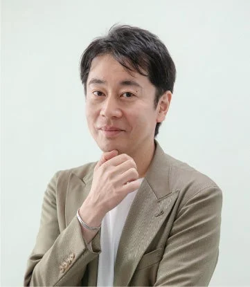薄茶色のジャケットを着た日本人男性が、顎に手を当ててカメラを見つめているポートレート写真。真面目な表情で何かを考えているような様子が伺えます。ビジネスシーンやカジュアルな場面にも合う服装です。