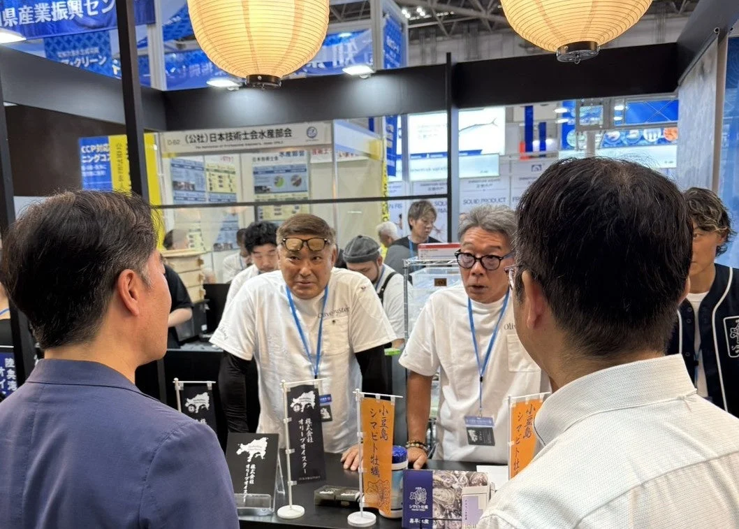 展示会場のような場所で、複数の人々がブースで交流している様子。白いTシャツを着た2人の男性が来場者と話しており、ブースには「株式会社オリーブオイスター」や「小豆島シマアビ」といった表示が見られる。天井からは和風の提灯が吊るされており、水産関連の展示会と推測される。