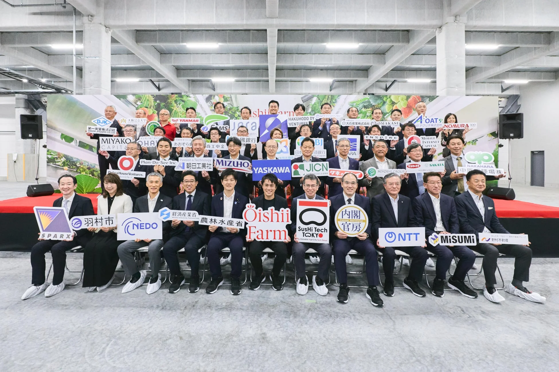 多数の企業、政府機関、大学などの代表者が一堂に会し、それぞれの組織名が書かれたボードを掲げて記念撮影をしている。