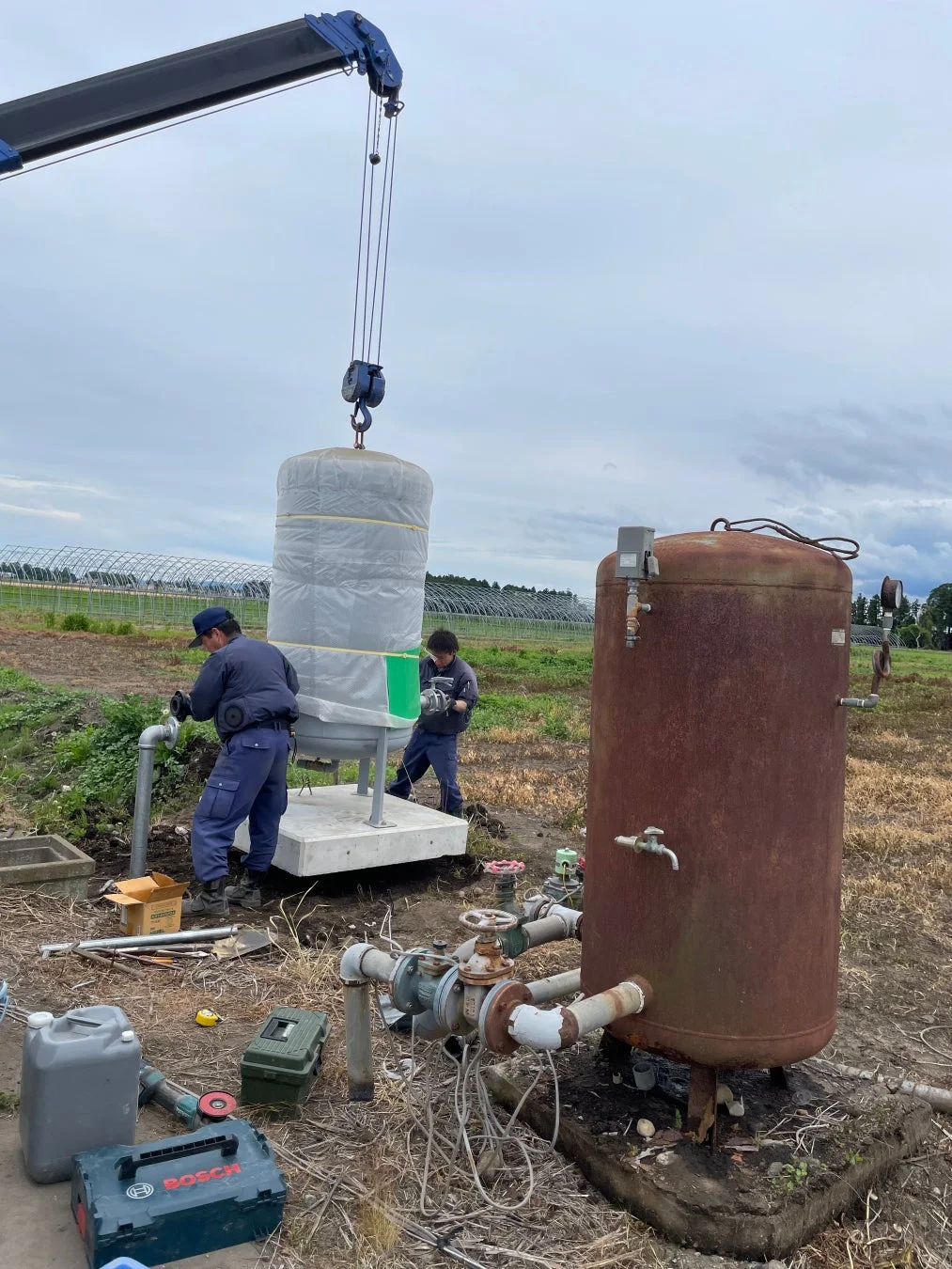農地で2人の作業員がクレーンを使い、新しい水処理タンクを設置している様子。