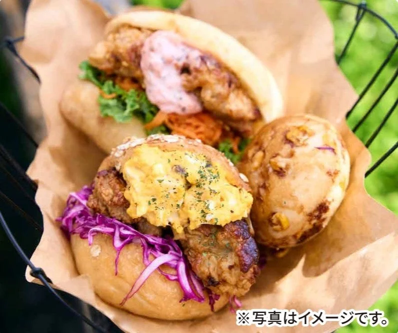 バスケットに盛られた2種類のフライドチキンサンドと、丸いパンが写っています。手前のサンドは紫キャベツと卵ソースで彩られ、奥のサンドはレタスとピンクのソースが特徴的です。屋外で楽しむ美味しそうな軽食の様子を捉えています。