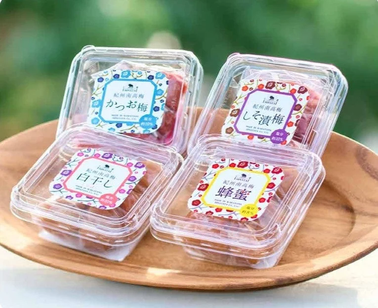 木製の皿に紀州南高梅の梅干しが4種類並んでいます。かつお梅、しそ漬梅、白干し、蜂蜜梅が小分けパックに入っており、それぞれ塩分濃度が記載されています。贈り物や食べ比べに最適な商品です。