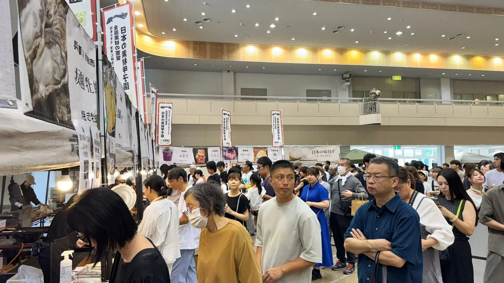 多くの人々で賑わう屋内イベント会場の様子。日本の素材甲子園と書かれた旗が多数見られ、全国各地の食材や焼きたて、スムージーなどの食品ブースが並んでいる。来場者は様々な商品を見て回っている。