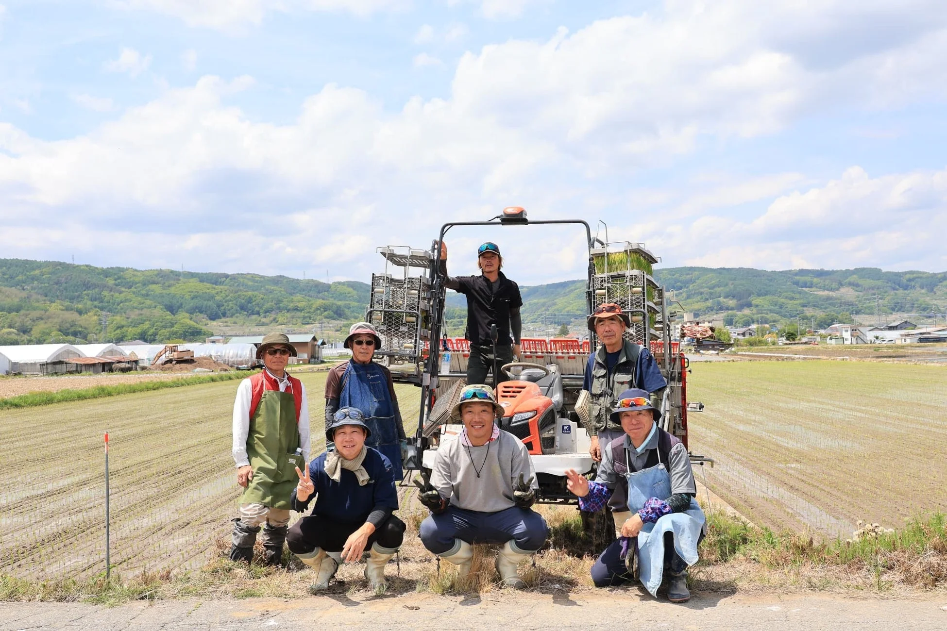 水田を背景に、田植え機と共に7人の人々が笑顔でポーズをとっています