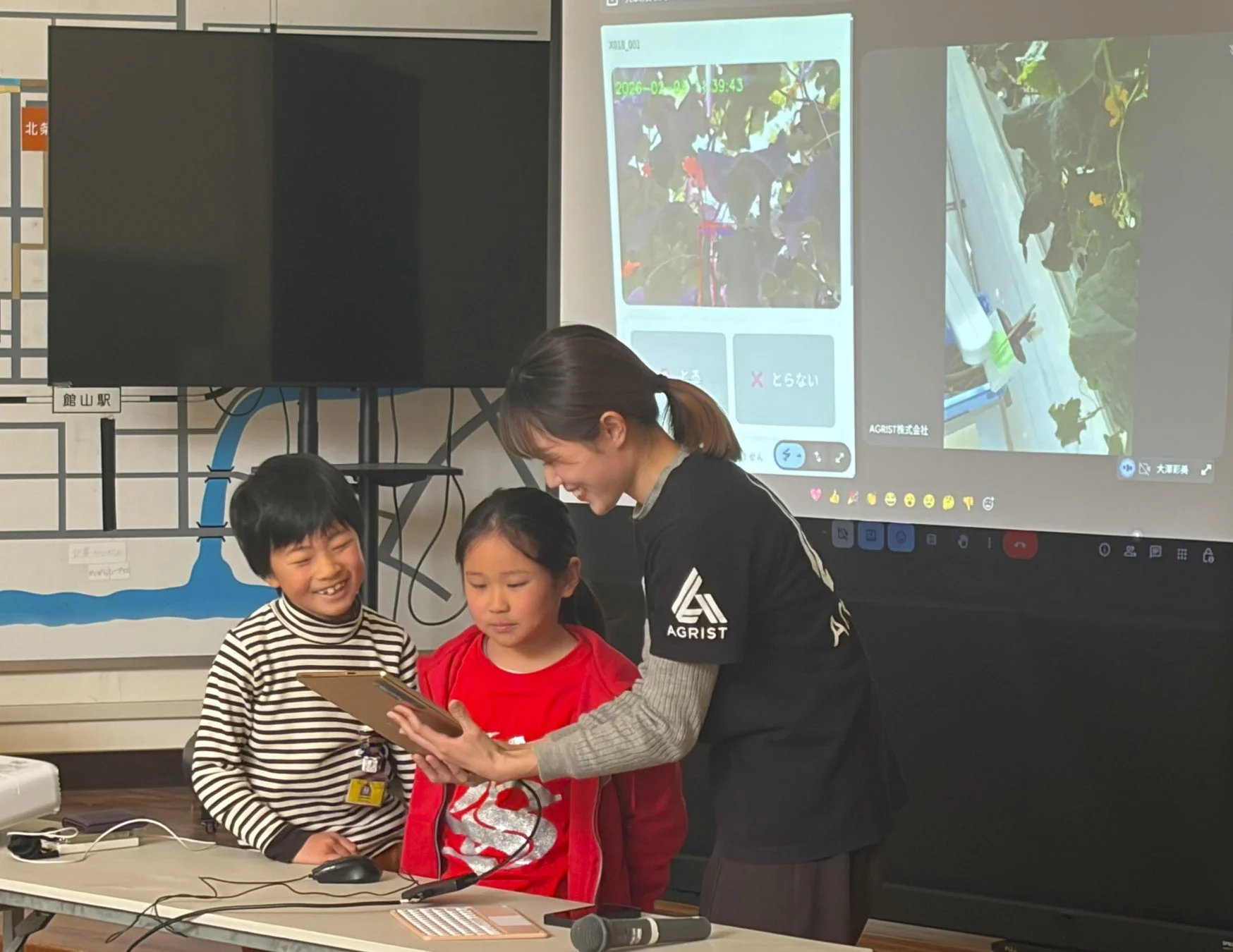 女性が子供たちにタブレットを使って説明している様子。背景のスクリーンにはAGRIST株式会社の農業技術に関する映像