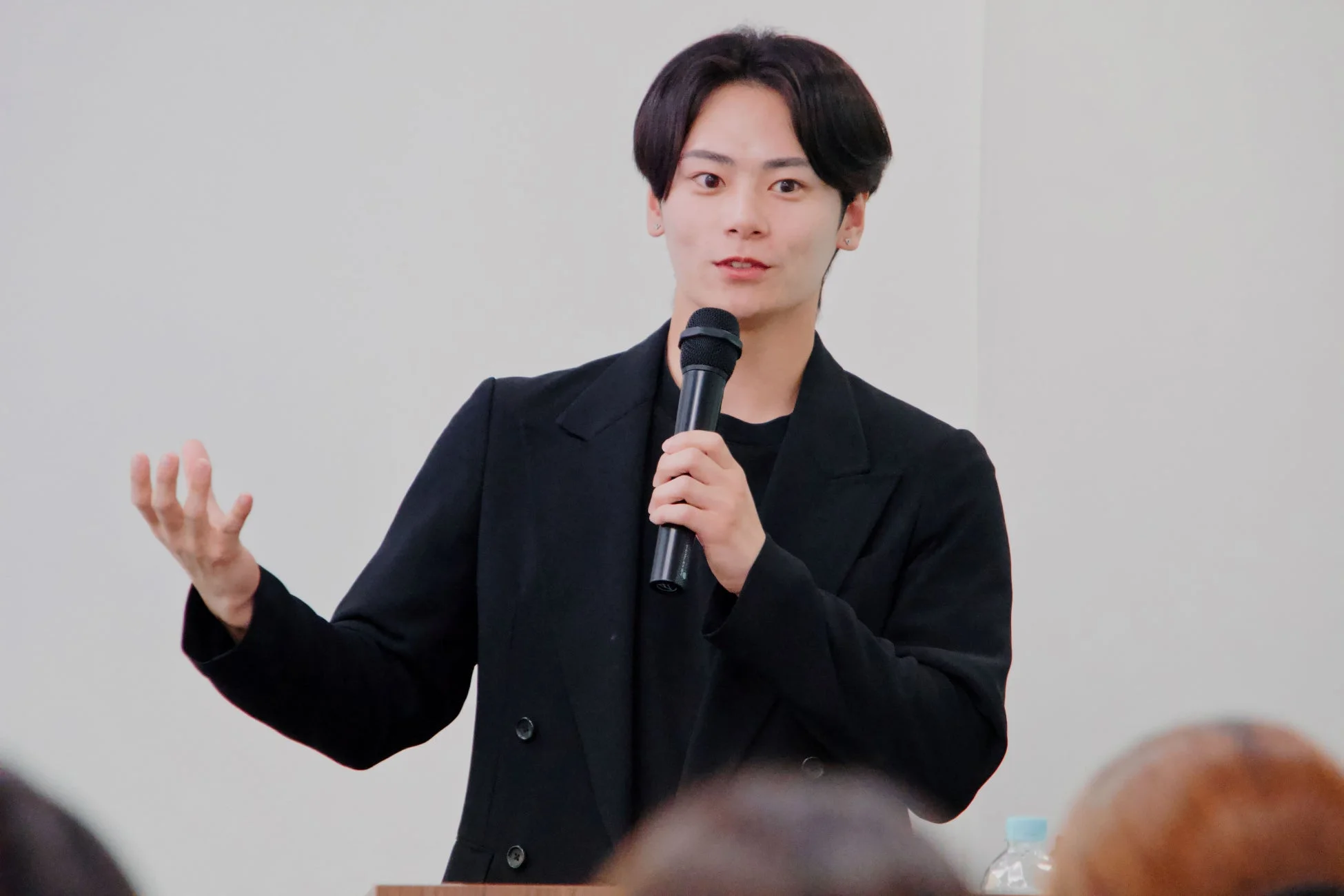 黒いジャケットを着た若い男性が、マイクを片手に身振り手振りでスピーチやプレゼンテーションを行っています