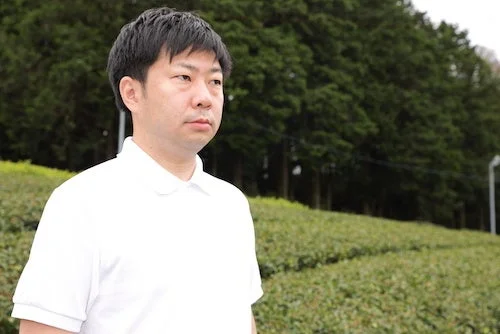 白いポロシャツを着た日本人男性が、緑豊かな茶畑と木々を背景に立っています。穏やかな表情で少し右方向を見ています。