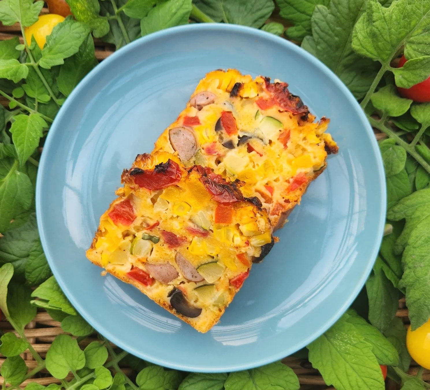青い皿に盛り付けられた、色とりどりの夏野菜とソーセージが入ったフリッタータまたはキッシュ。周囲には緑の葉とミニトマトが添えられ、新鮮で彩り豊かな食卓を演出しています。