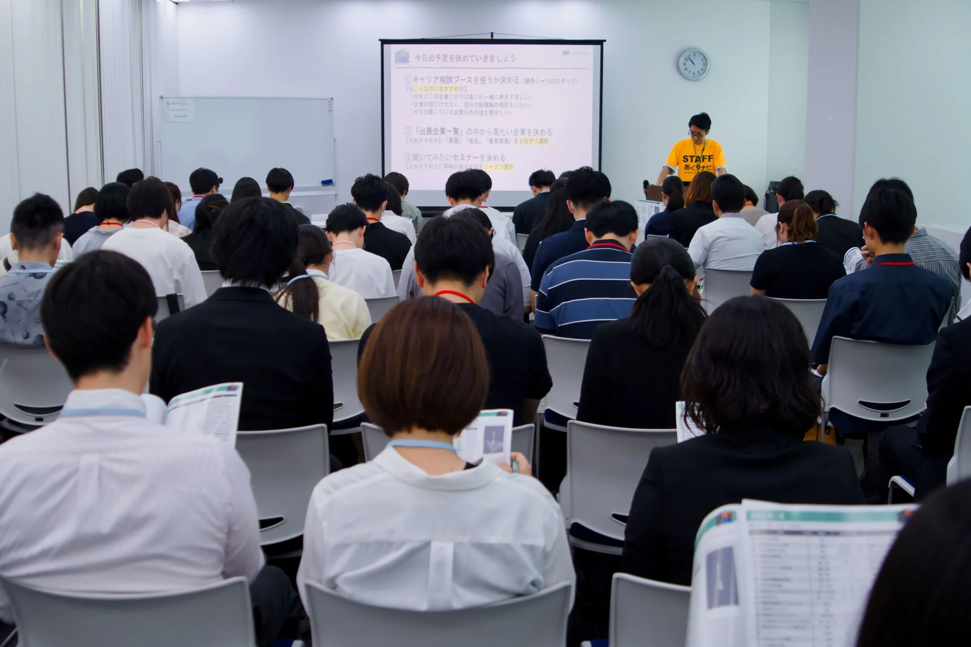 プロジェクターに今日の予定が表示された会場で、多くの参加者が熱心に講演を聞いている様子